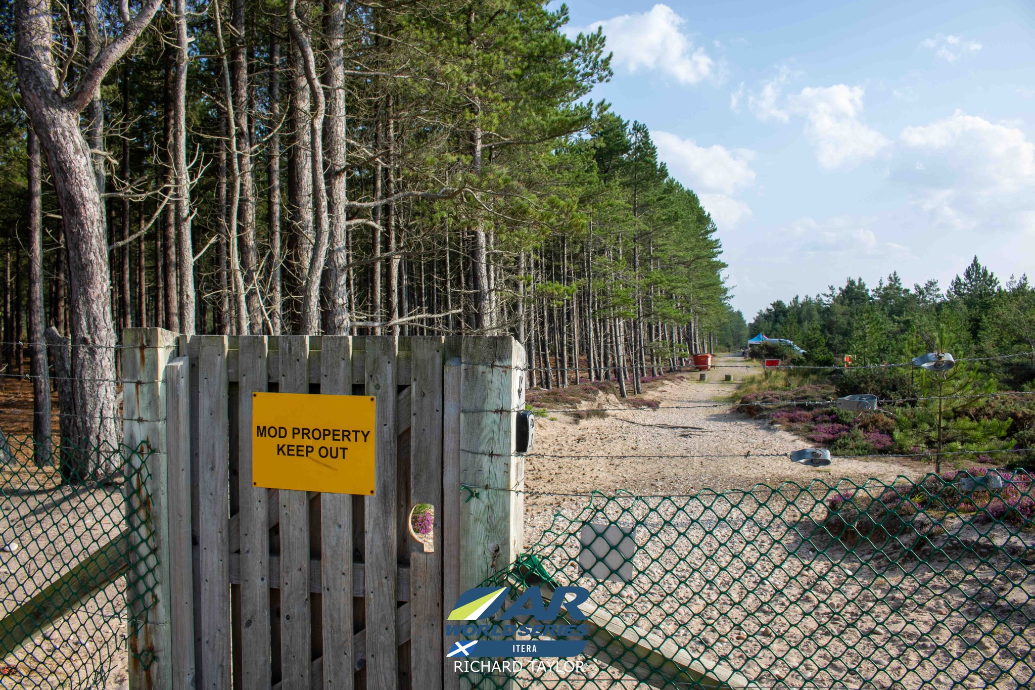 Small wooden gate set in a barbed wire fence with a yellow sign reading 'MOD Property Keep Out'. On the other side you can just see the small gazebo where we got instructions.